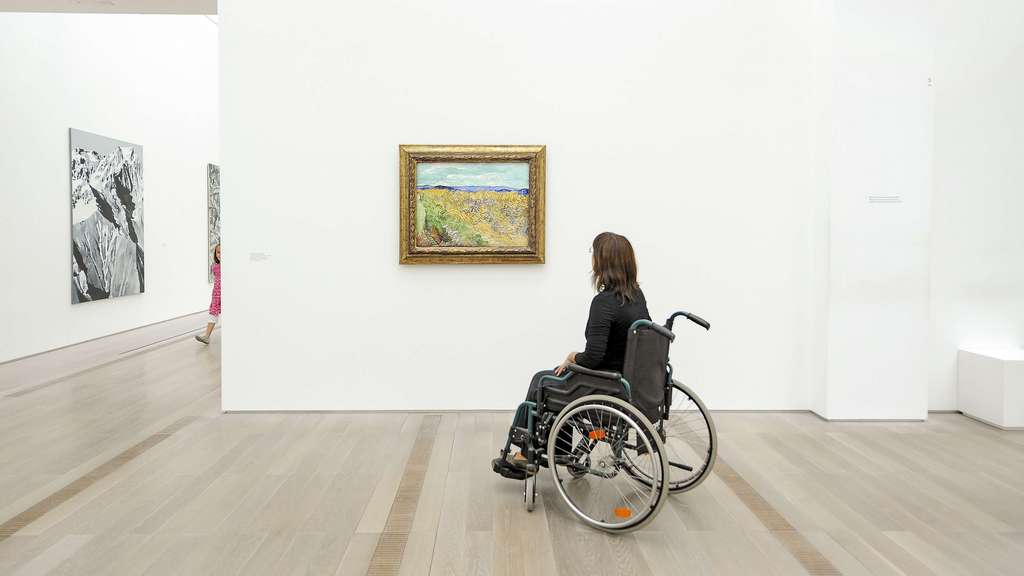 A person in a wheelchair looks a painting of a lanscape in a museum hall