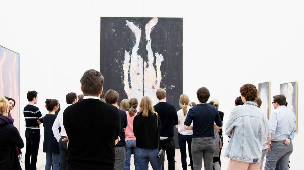 Large group of people look at a black painting on the wall with their backs to camera.