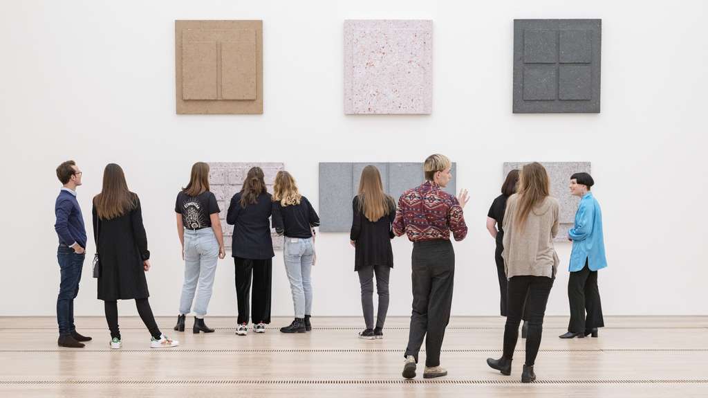 Des jeunes dans une salle d'exposition regardent un mur rempli d'œuvres d'art.