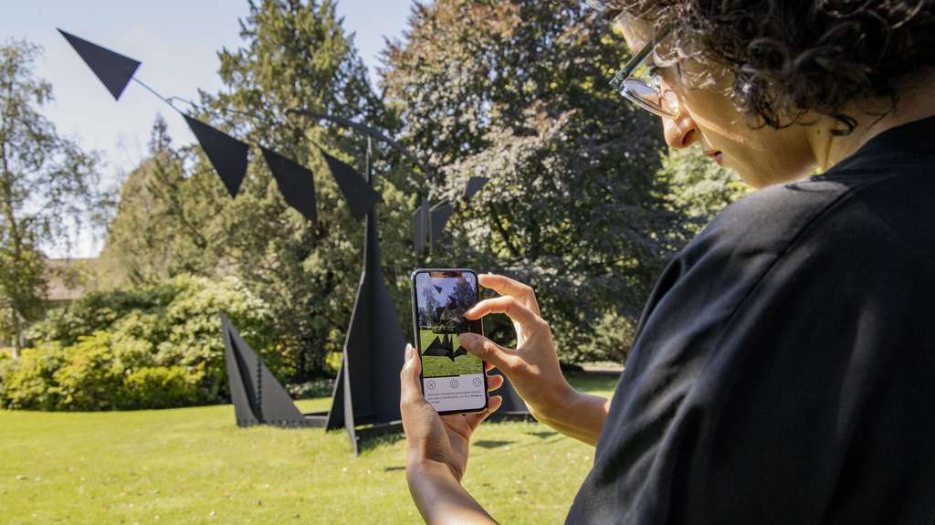 Person mit Smartphone in der Hand vor einem Skulptur in einem Park.