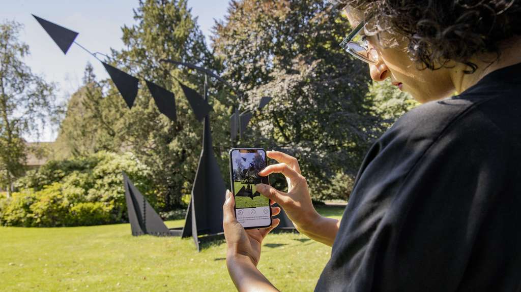 Personne avec un smartphone à la main devant une sculpture de E. Kelly dans un parc.