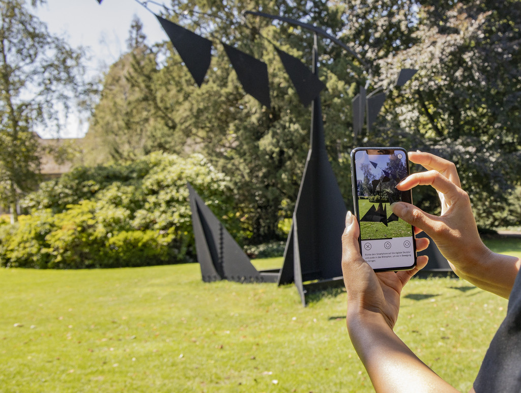 Personne avec un smartphone à la main devant une sculpture de E. Kelly dans un parc.
