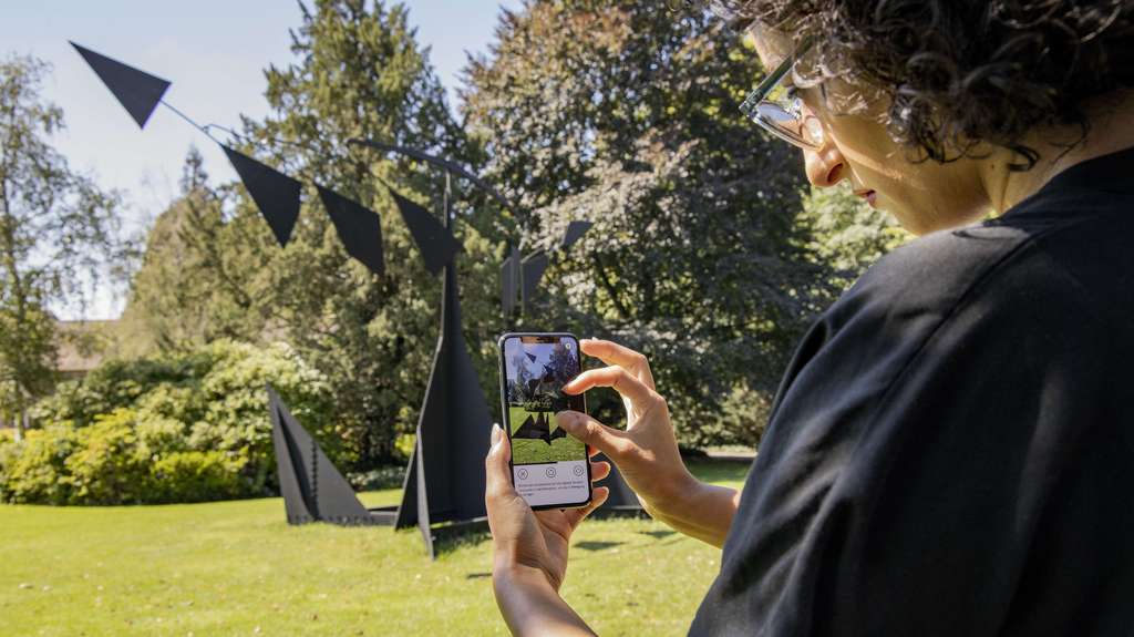 Woman with a smartphone in her hand in front of Kelly sculpture in park.