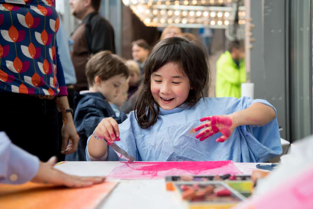 A happy young girl plays with pink colour at a table in a museum studio room.