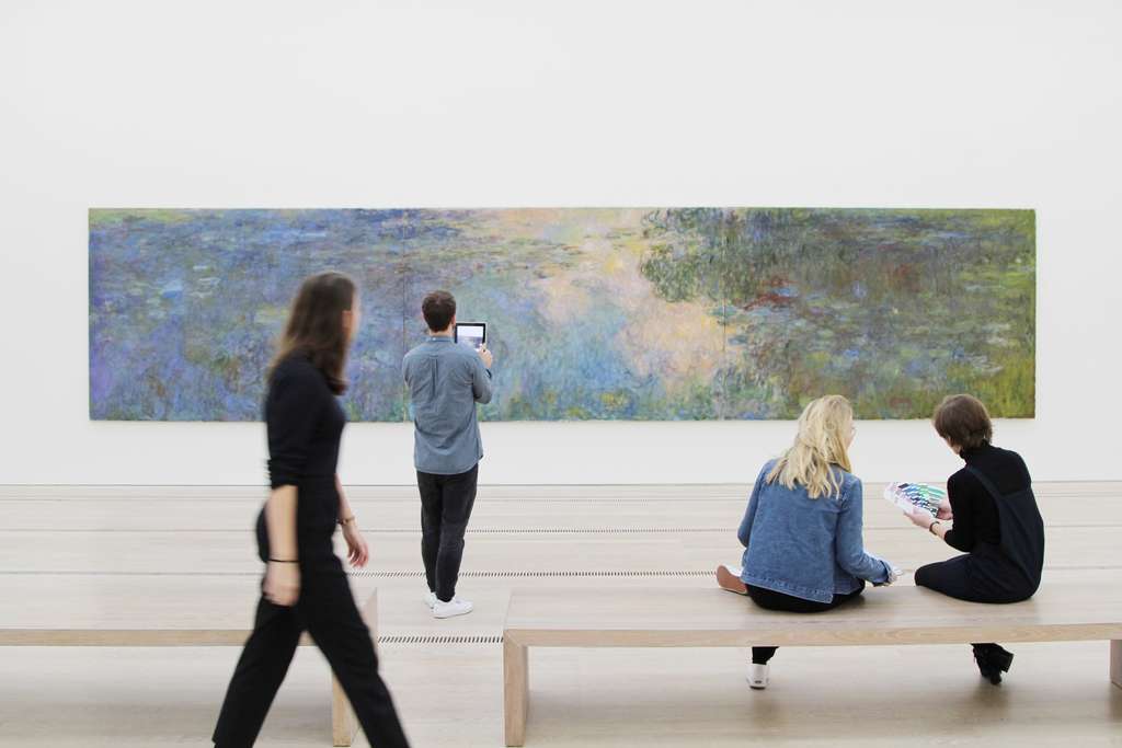 Visitors stand and sit in front of a painting by Claude Monet.