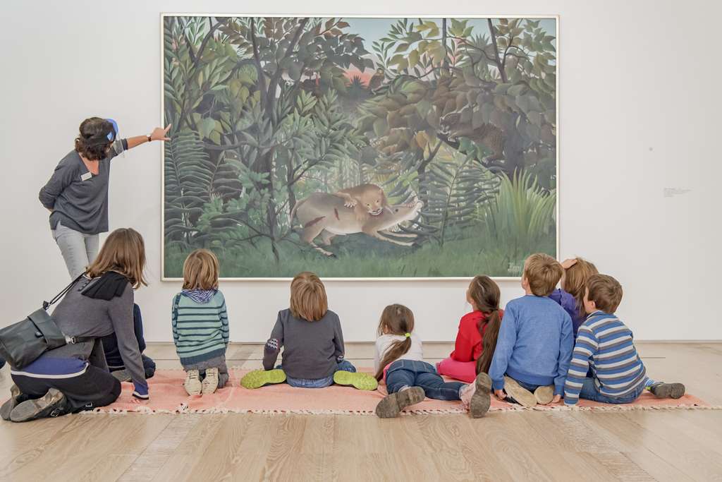 A woman presents a work of art by Rousseau to a group of children.